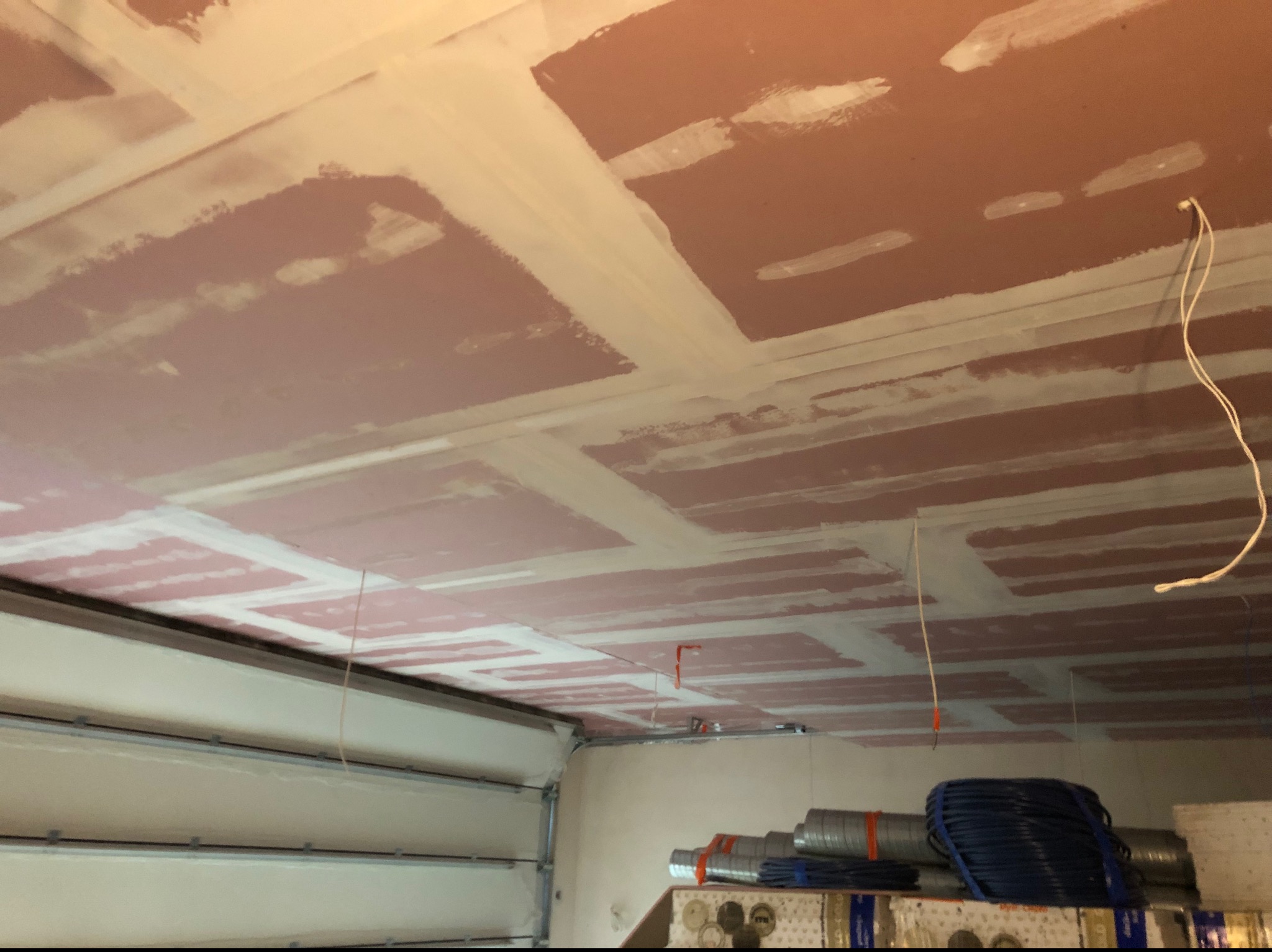 Różowe płyty karton-gips na suficie garażu, z widocznymi łączeniami szpachlowanymi na biało. W tle widoczne materiały budowlane i brama garażowa.