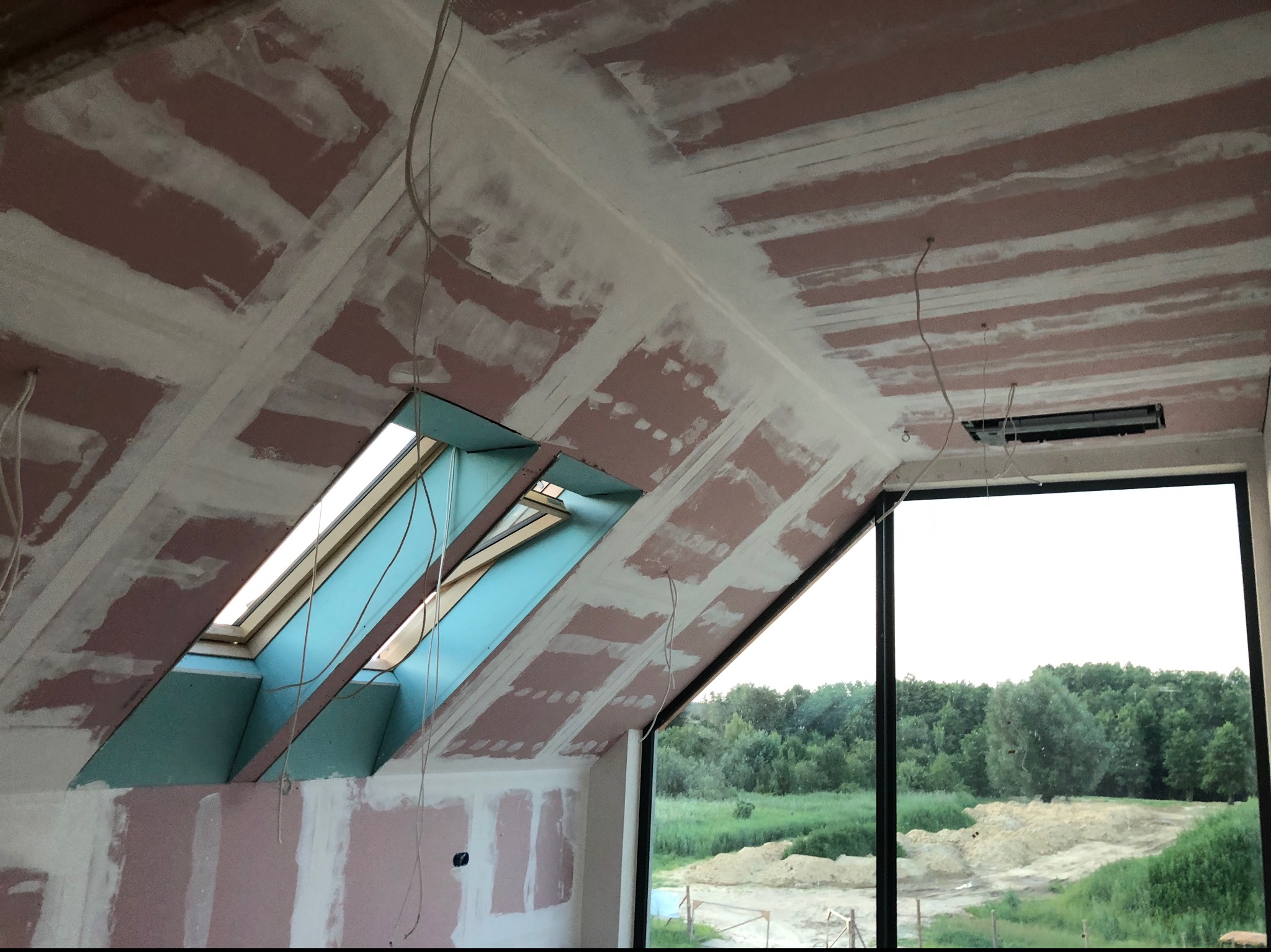 Poddasze z oknem dachowym w trakcie wykańczania: płyty karton-gips na skosach, wstępnie szpachlowane łączenia, widoczne przewody elektryczne. W tle zieleń za oknem.