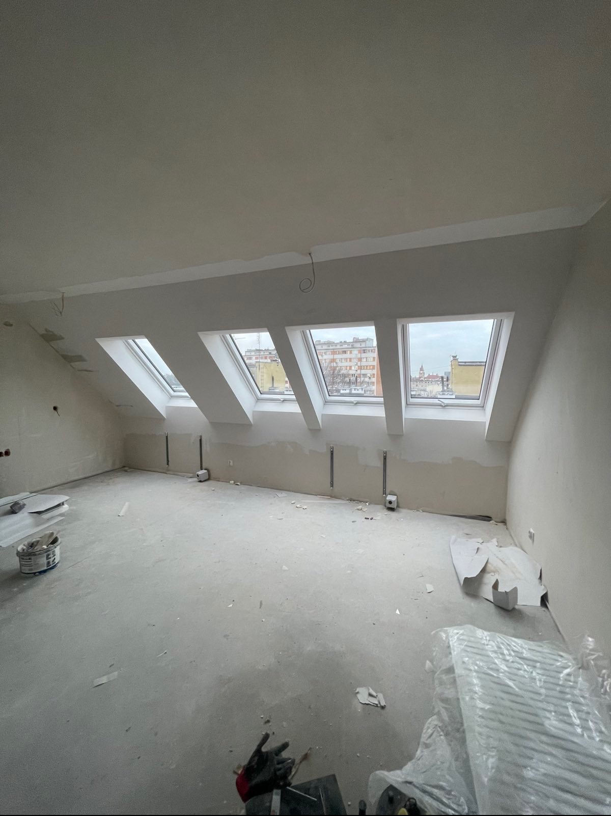 Poddasze po szpachlowaniu, widoczne trzy okna dachowe z widokiem na miasto. Ściany przygotowane do malowania, na podłodze resztki materiałów budowlanych i narzędzia.