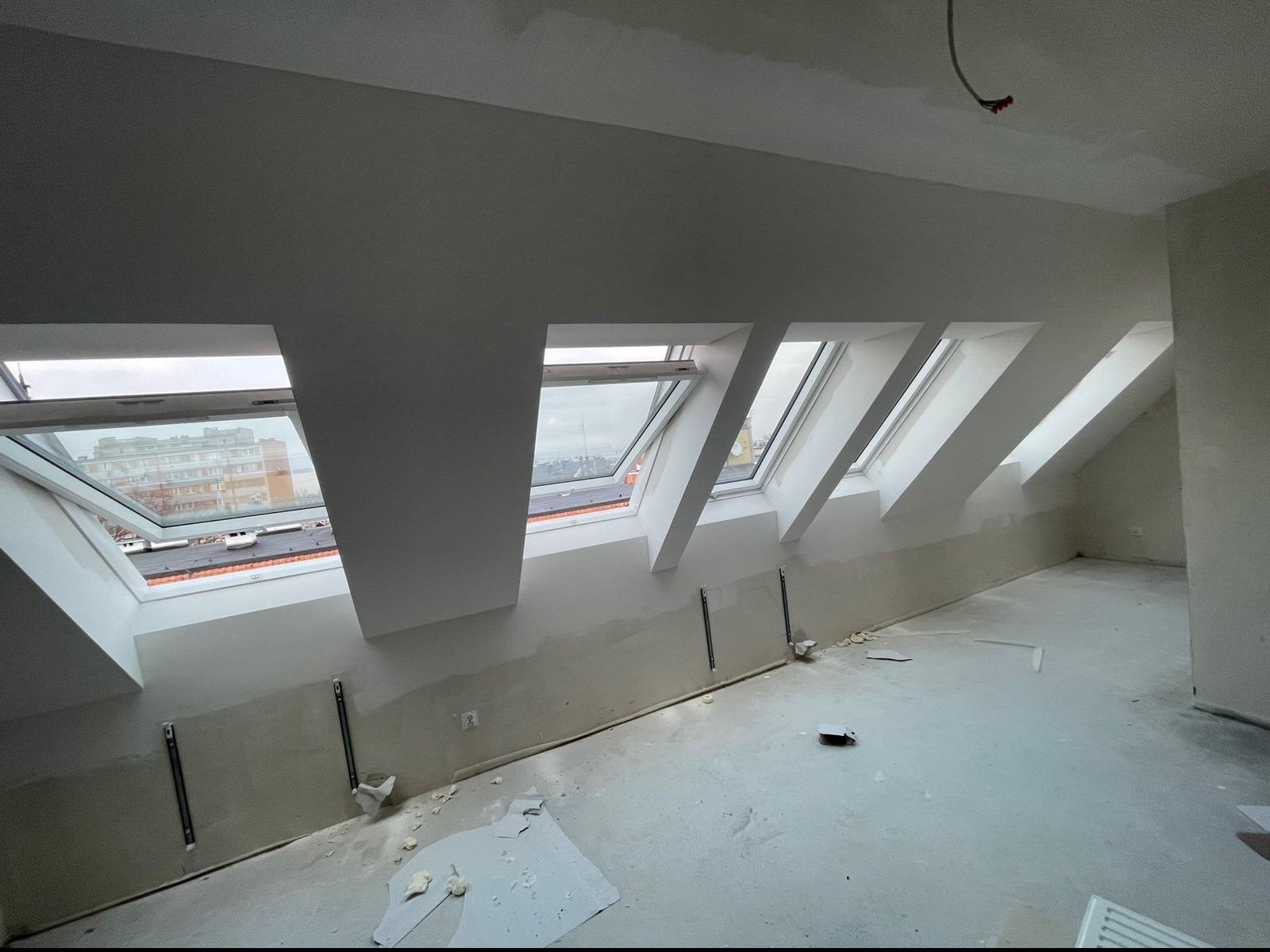 Poddasze w trakcie prac wykończeniowych: widoczne okna dachowe, ściany przygotowane do malowania, surowa podłoga. Elementy montażowe na ścianach.