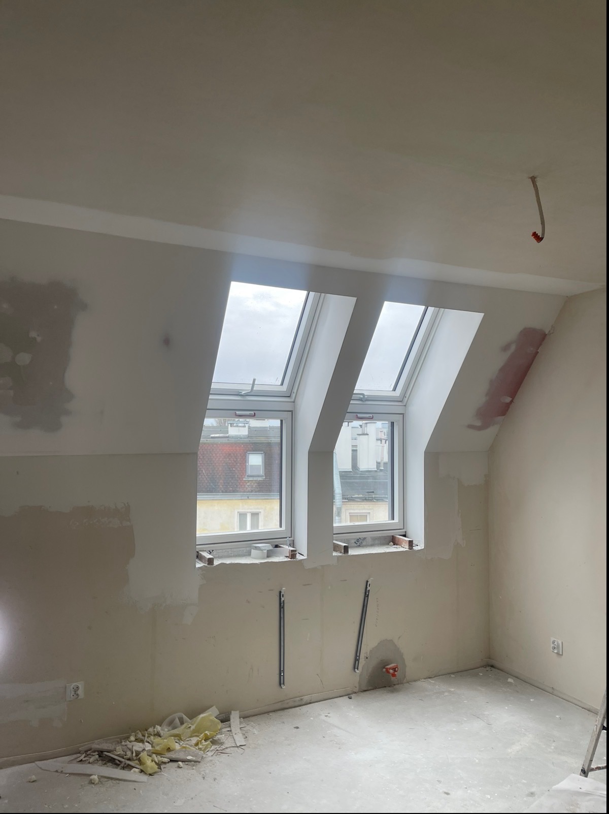 Pokój na poddaszu w trakcie remontu. Widoczne okna dachowe, ściany w trakcie szpachlowania, fragment instalacji elektrycznej i resztki materiałów budowlanych na podłodze.