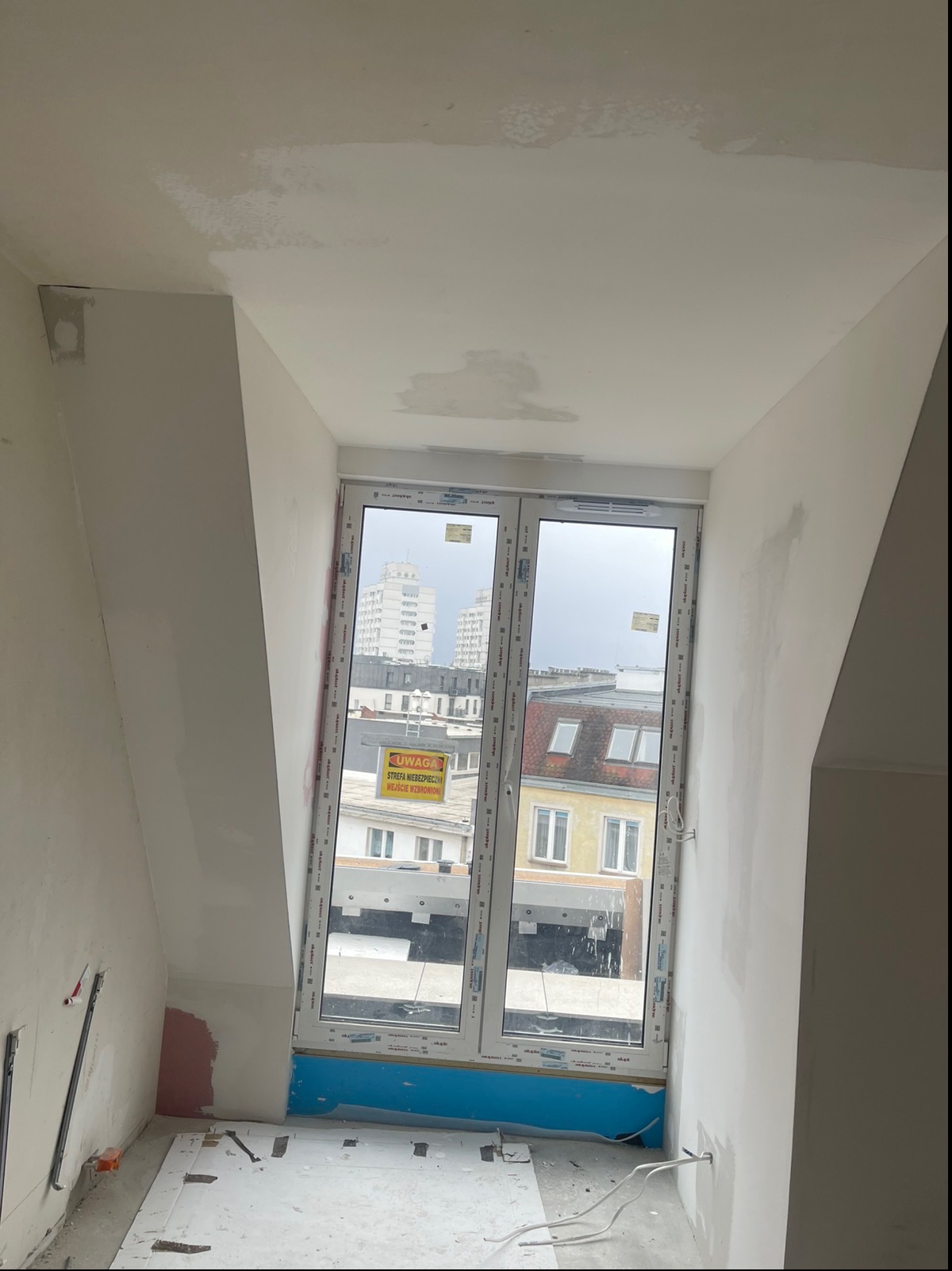 Poddasze w trakcie wykańczania: okno z widokiem na miasto, ściany częściowo pokryte gładzią, widoczne niedoskonałości i przygotowania do dalszych prac wykończeniowych.