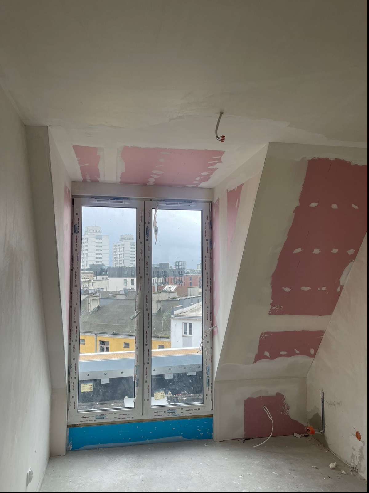 Poddasze w trakcie prac wykończeniowych: szpachlowane ściany z widocznymi fragmentami płyt karton-gips, okno z widokiem na miasto, przygotowane instalacje elektryczne.