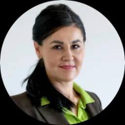 Portret uśmiechniętej kobiety z ciemnymi włosami w zielonej koszuli i brązowej marynarce, na jasnym tle.