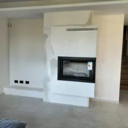 Nowoczesny wkład kominkowy z czarną ramą, w surowej, białej obudowie z gładzią, widoczne naklejki informacyjne na szybie, wnętrze wyłożone szarym materiałem.