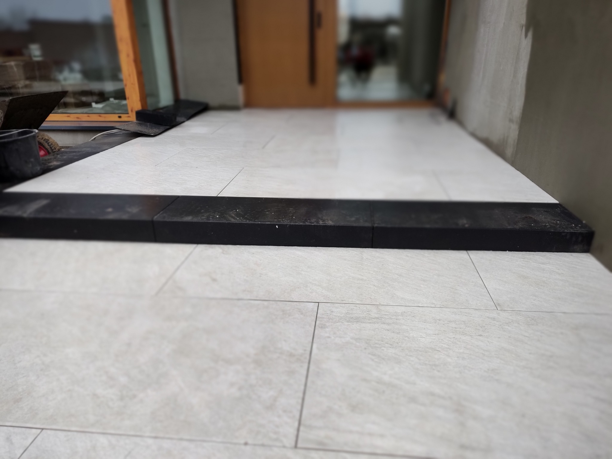 Wyłożony płytkami jasny taras przed wejściem do domu z widocznym czarnym progiem i fragmentem narzędzi brukarza.