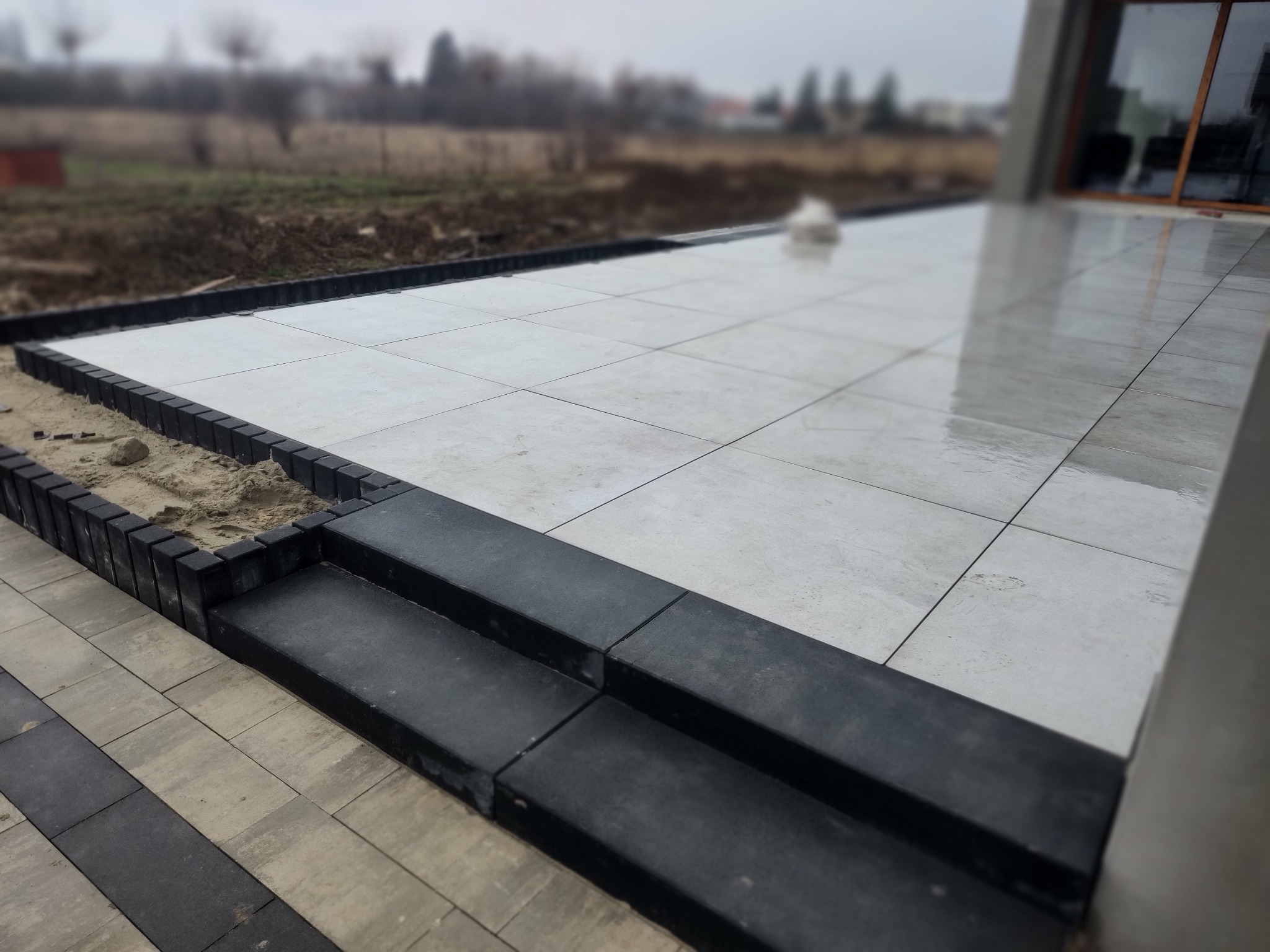 Jasnoszara, kwadratowa nawierzchnia z płytek na zewnątrz budynku, z czarnymi obrzeżami i dwustopniowymi schodami z ciemnych elementów.