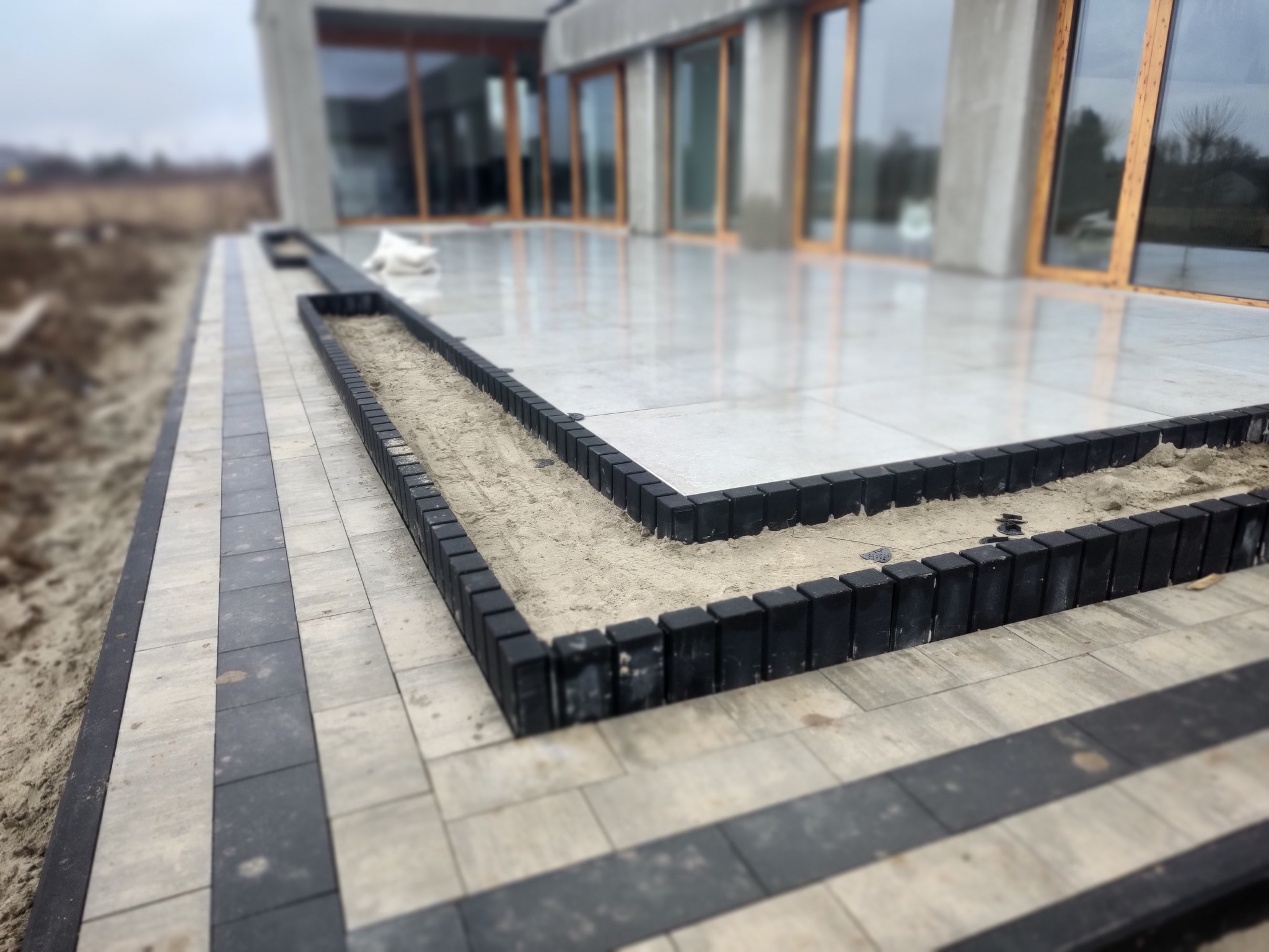 Układanie jasnoszarej kostki brukowej z ciemnymi pasami, obrzeża z czarnych kostek, przygotowane miejsce na nasadzenia roślin, nowoczesny dom w tle.