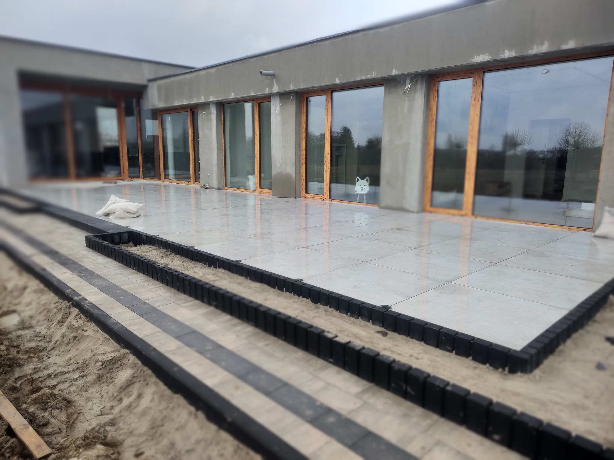 Montaż obrzeży z kostki brukowej na tarasie z płyt ceramicznych, widoczne przygotowane podłoże z piasku, duże okna tarasowe w tle.