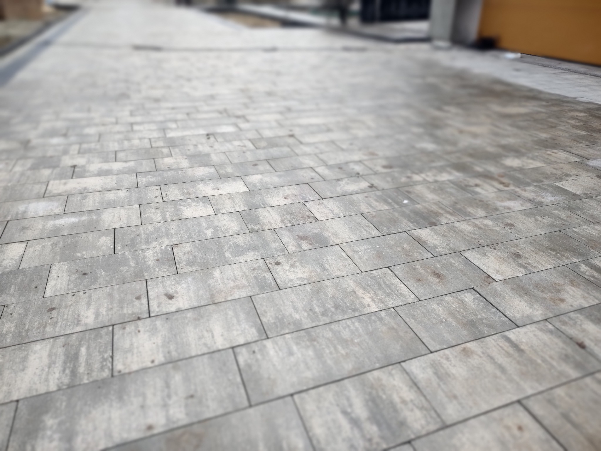 Szeroki chodnik wyłożony szarą, prostokątną kostką brukową w perspektywie zbieżnej, z rozmytym tłem sugerującym otoczenie miejskie.