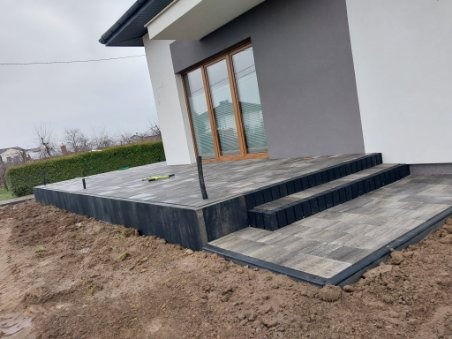 Nowoczesny taras z kostki brukowej w odcieniach szarości, z trzema stopniami prowadzącymi do domu, widoczne elementy wykończeniowe i niezagospodarowany teren wokół.