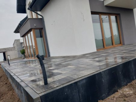 Nowoczesny taras z szarej kostki brukowej ułożonej wzorem jodełki, widok na elewację domu z dużymi oknami przesuwnymi.