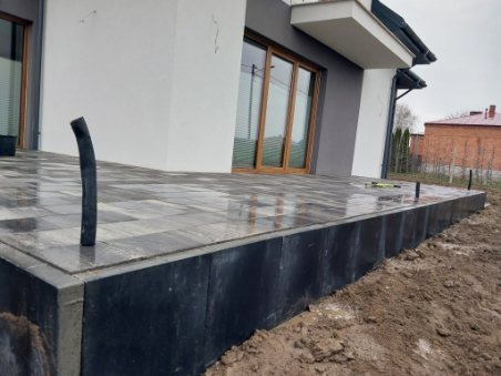 Nowoczesny taras z szarej kostki brukowej ułożonej na podwyższeniu z czarnych paneli, widok na dom z brązowymi oknami i drzwi balkonowymi, w tle ceglany budynek.