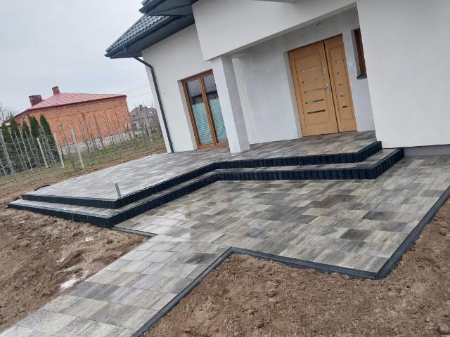 Nowoczesny podjazd i schody wejściowe wyłożone szaro-brązową kostką brukową, z czarnym obrzeżem, prowadzące do jasnego domu z drewnianymi drzwiami.