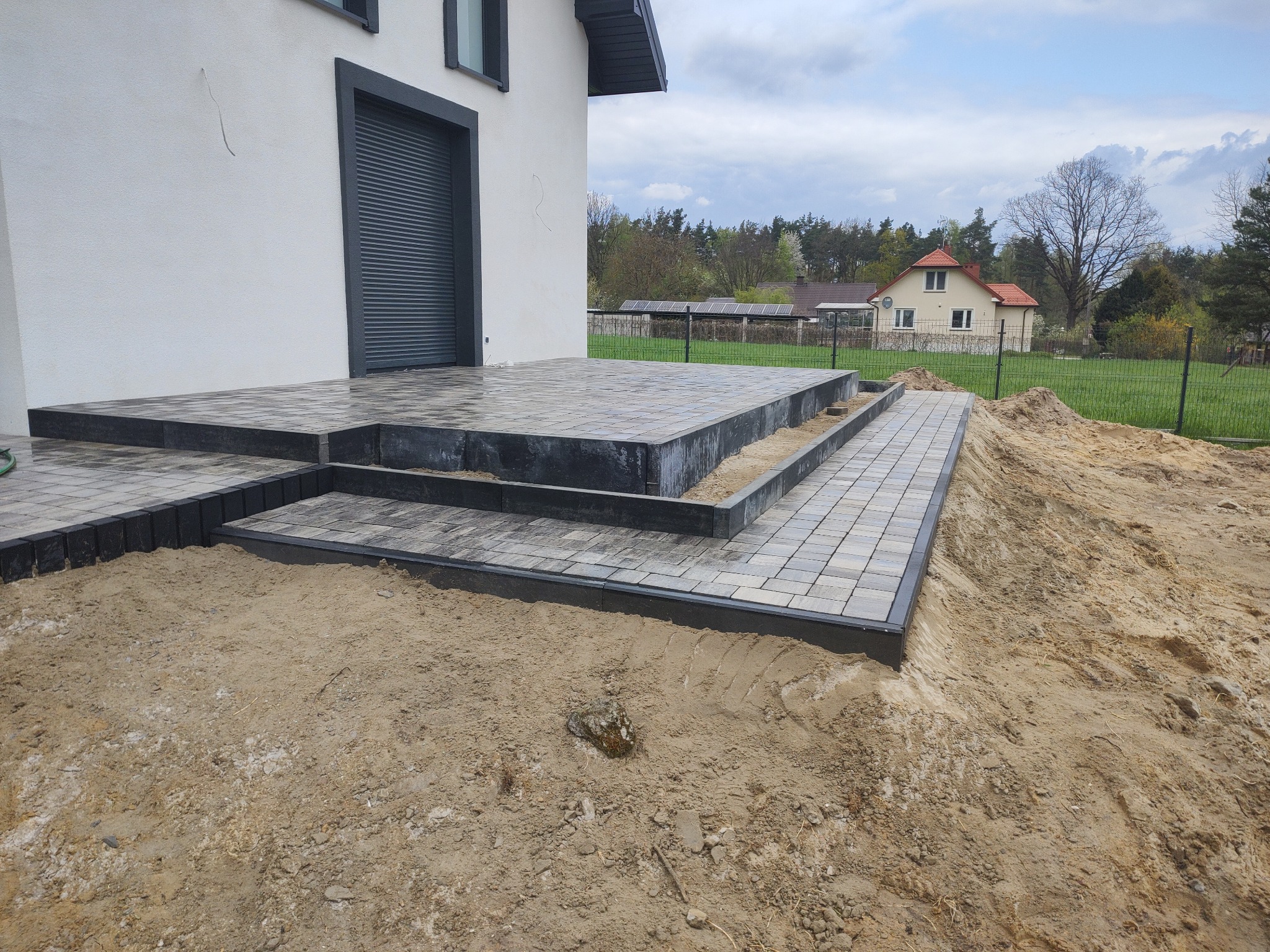 Ułożona kostka brukowa tworząca schody i chodnik przy białym domu z szarymi elementami, w tle trawnik, drzewa i dom z czerwonym dachem.