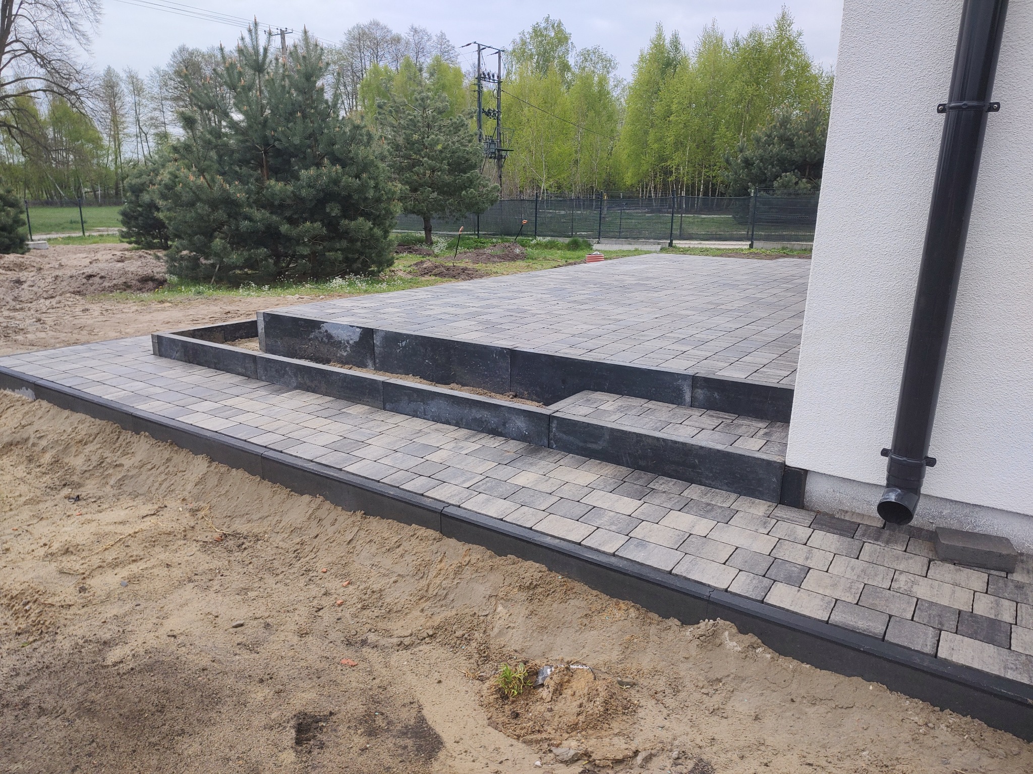 Nowo ułożony taras z kostki brukowej z trzema stopniami, obrzeżami i rurą spustową przy białej ścianie domu, widok na ogród w tle.