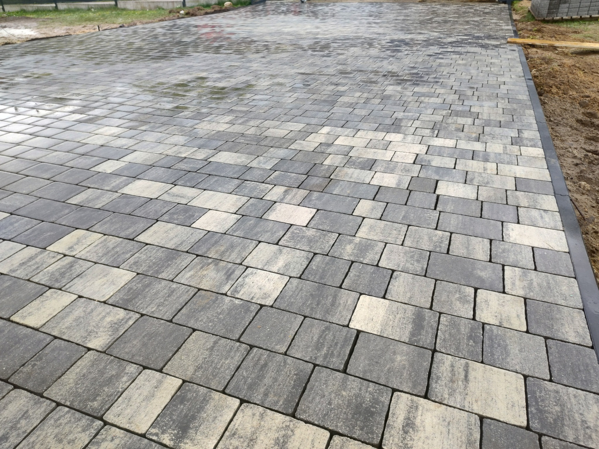 Świeżo ułożona, częściowo mokra nawierzchnia z kostki brukowej w odcieniach szarości i beżu, ułożona w regularny wzór, widoczne obrzeża i przygotowany teren pod dalsze prace.