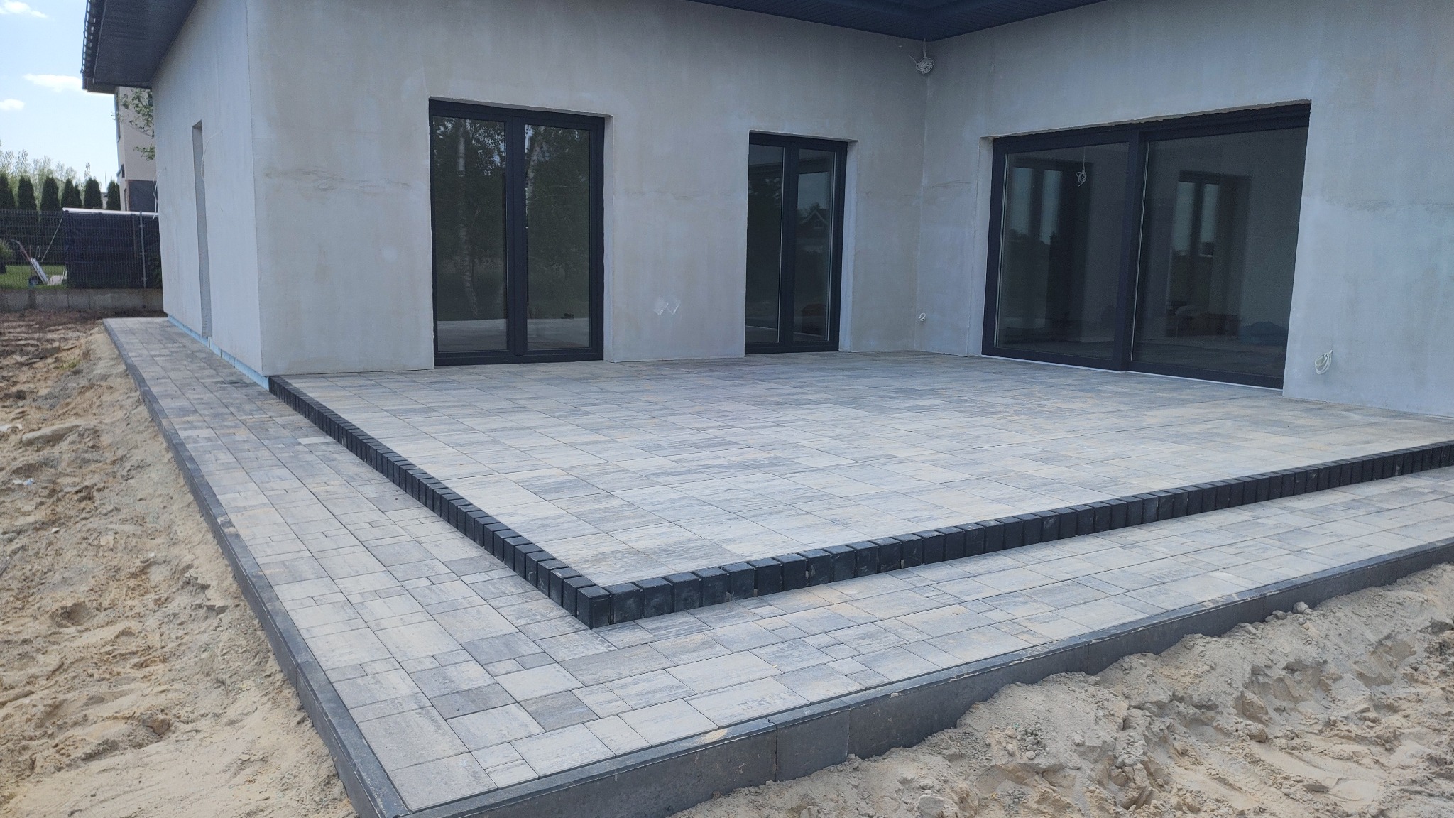 Nowoczesny taras z szarej kostki brukowej, wykończony ciemnymi obrzeżami, przylegający do surowego budynku z dużymi, ciemnymi oknami, otoczony piaskiem.