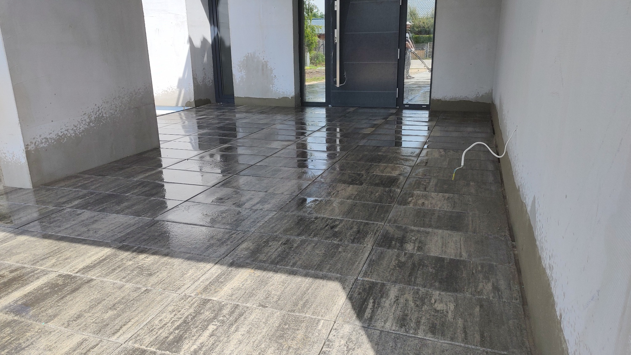 Świeżo ułożona, mokra, grafitowa kostka brukowa na tarasie przed wejściem do domu w trakcie prac wykończeniowych, widoczne niedociągnięcia na ścianach i wystający kabel.
