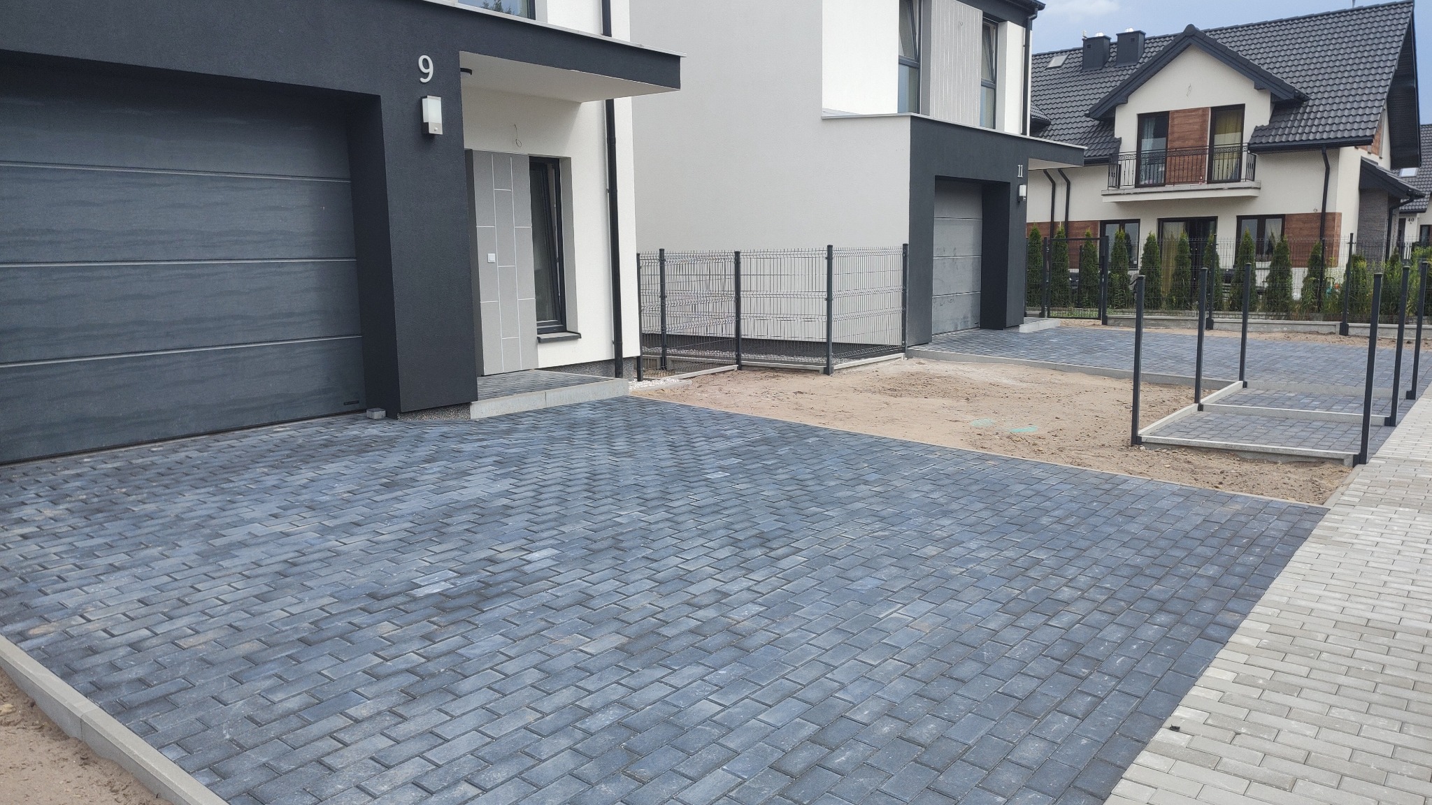 Ułożona szara kostka brukowa na podjeździe do nowoczesnych domów jednorodzinnych z garażami, widoczne obrzeża i przygotowane podłoże pod kolejne etapy prac.