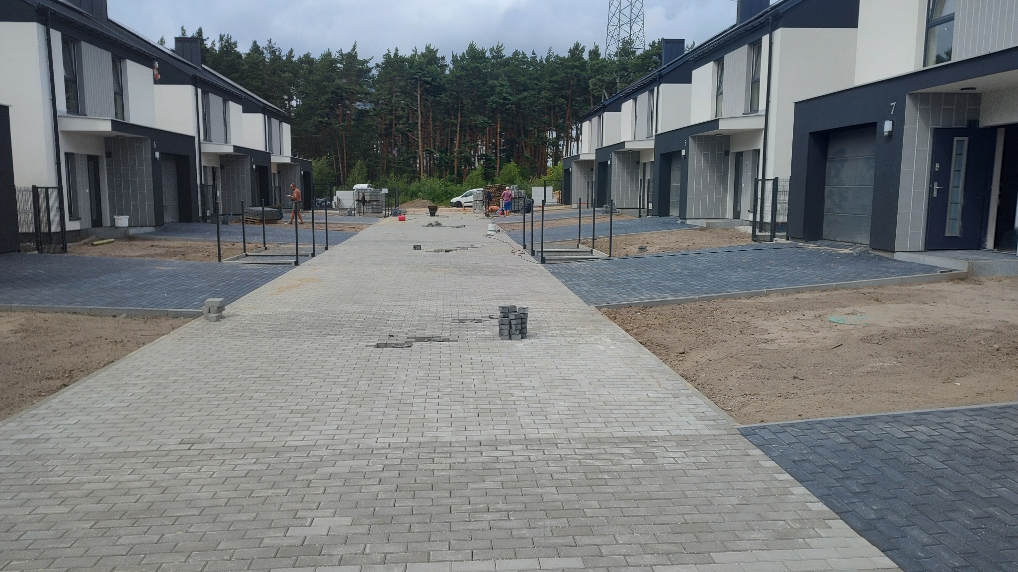 Ulica osiedlowa w trakcie budowy z jasnoszarej kostki brukowej, widoczne niedokończone fragmenty i sterta kostki, w tle szereg nowoczesnych domów.