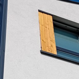 Pro Serwis Sp. z o.o. - Nowoczesne okno drewniane z żaluzjami i drewnianą osłoną, widok z dołu na tle błękitnego nieba.