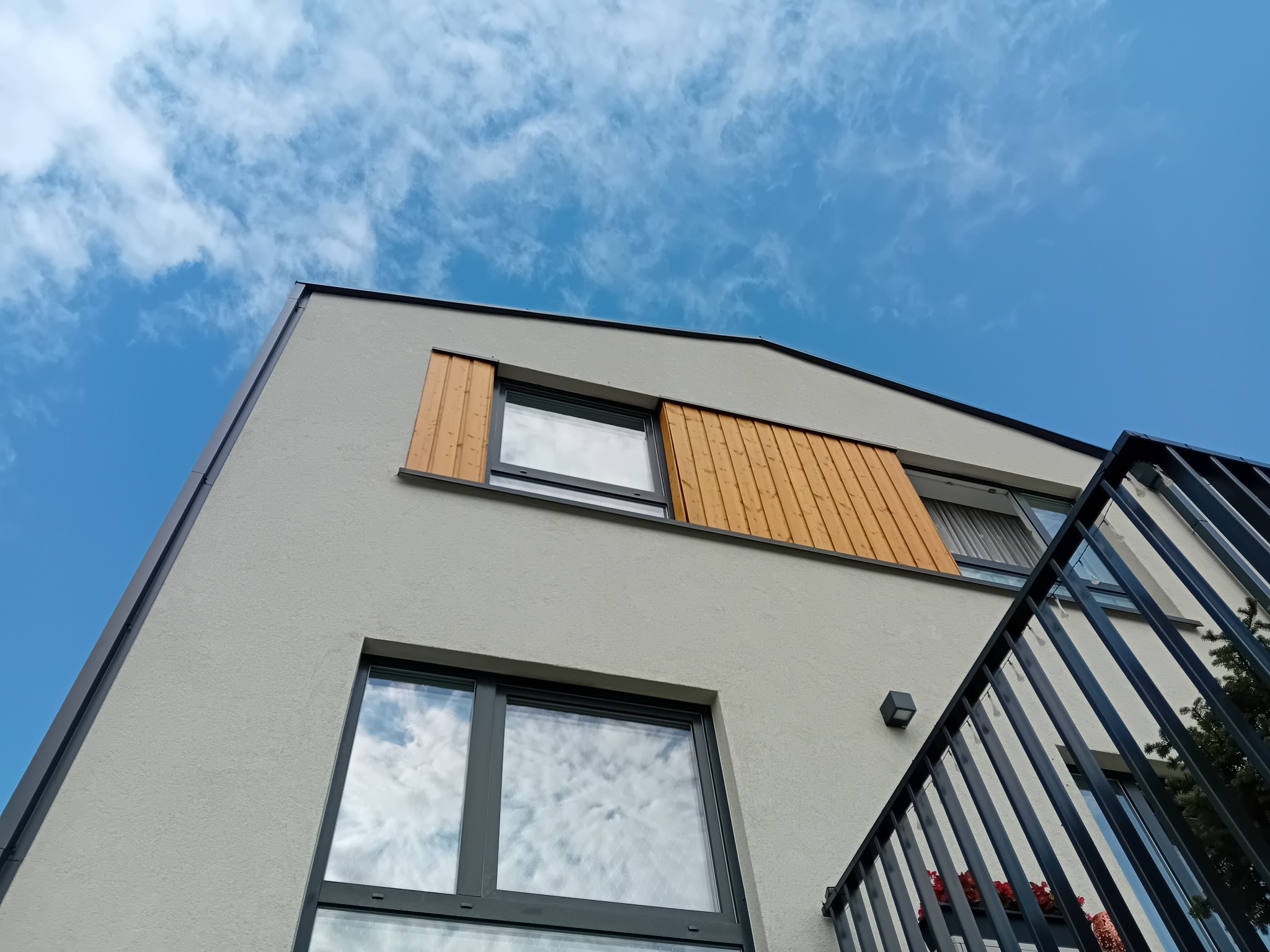 Nowoczesny dom z szarą elewacją, drewnianymi panelami i oknami z ciemnymi ramami, widok z dołu na tle błękitnego nieba z chmurami.