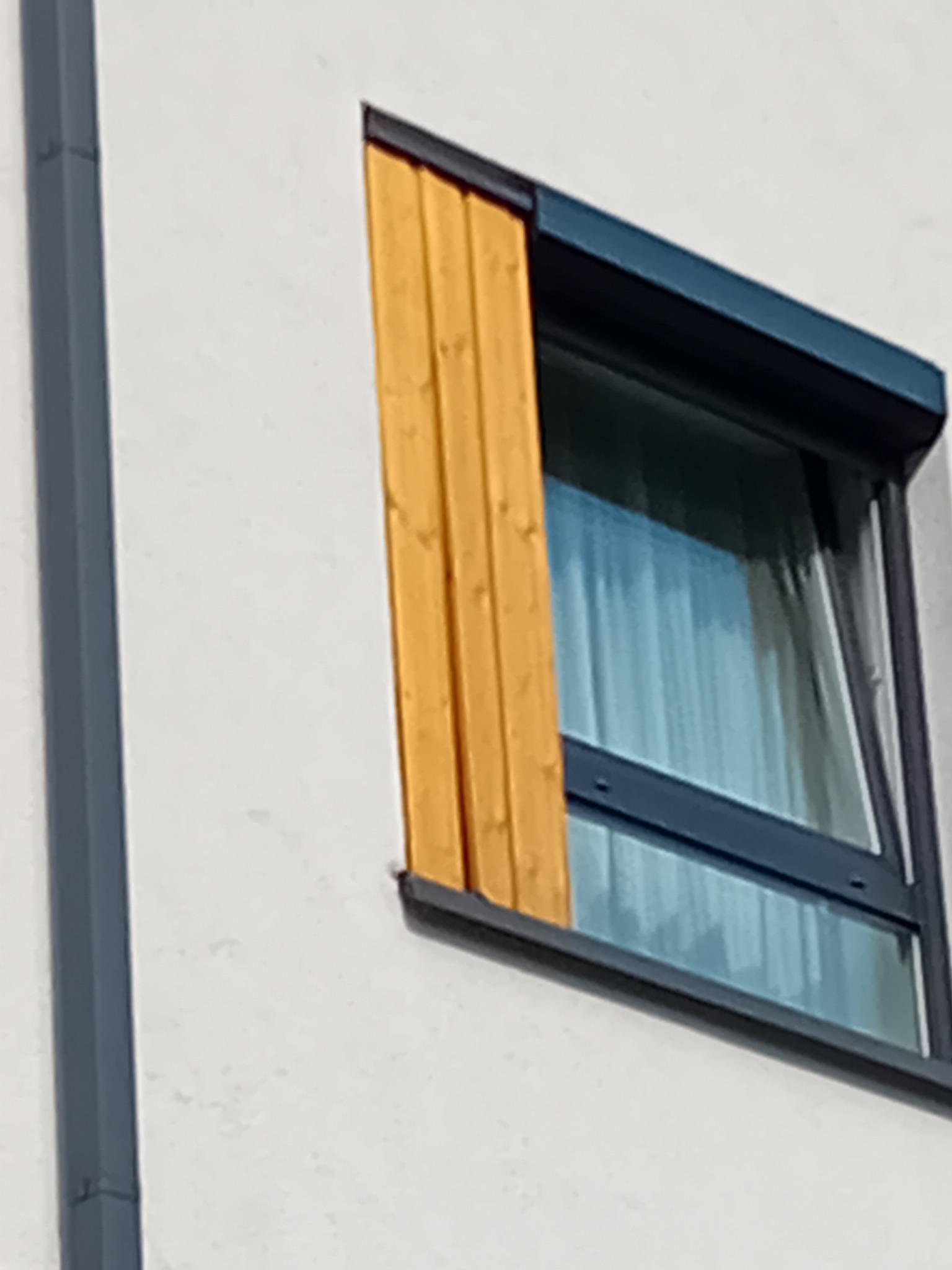 Fragment fasady z oknem z drewnianą listwą maskującą, widoczny fragment rynny w kolorze grafitowym.