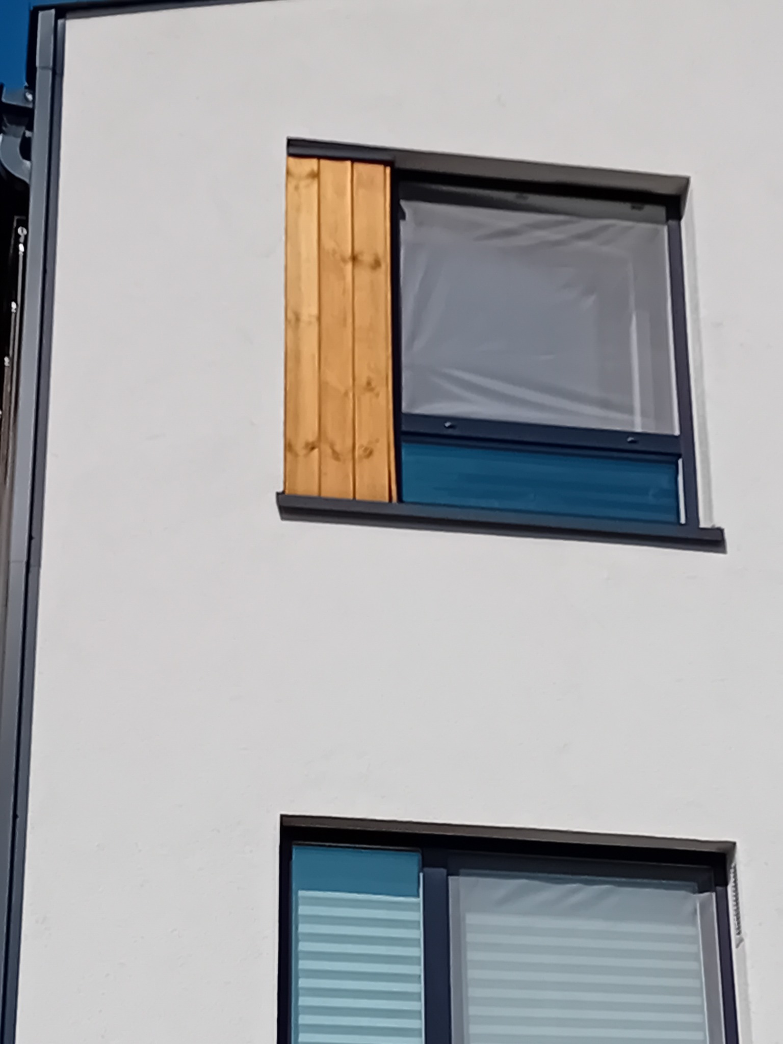 Fragment elewacji budynku z dwoma oknami: górne okno zabezpieczone folią ochronną i drewnianą listwą, dolne z żaluzjami poziomymi.