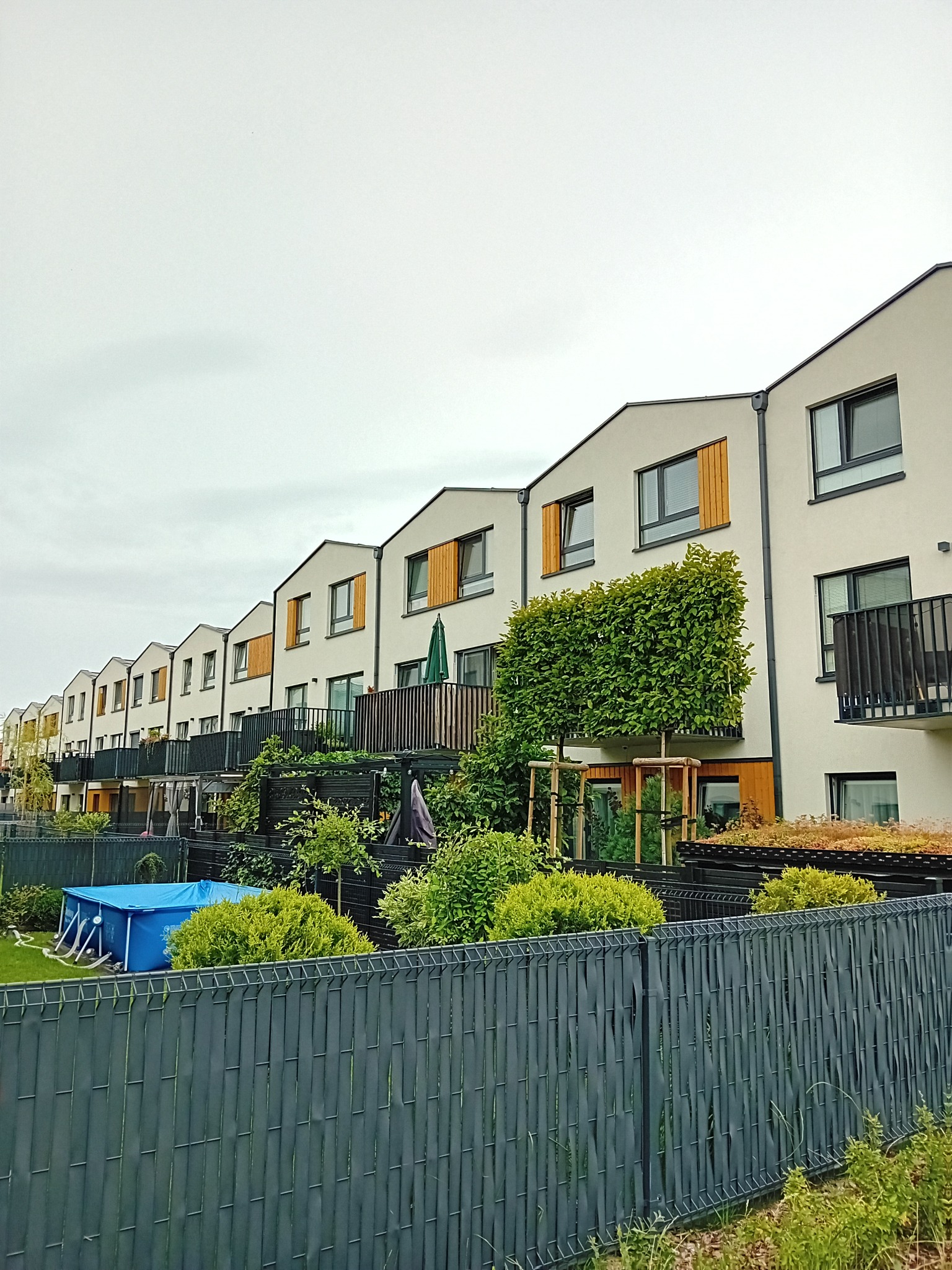 Szereg nowo wybudowanych, nowoczesnych domów szeregowych z drewnianymi okiennicami i balkonami, widoczny basen ogrodowy za ogrodzeniem.