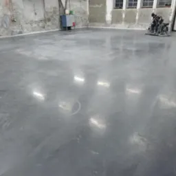 Operator w trakcie zacierania świeżo wylanej, szarej posadzki w dużym, surowym pomieszczeniu z oknami, widoczne refleksy świetlne na powierzchni.