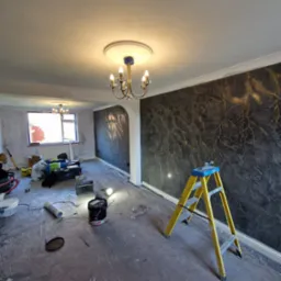 Pokój w trakcie remontu z dekoracyjną ścianą w odcieniach szarości i złota, żółta drabina, narzędzia malarskie i zabezpieczona folią podłoga.