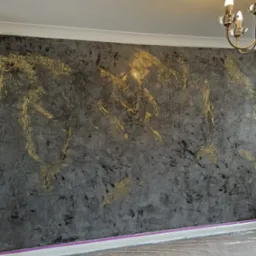 Ściana pokryta dekoracyjną szarą fakturą z nieregularnymi złotymi akcentami, widoczna listwa przypodłogowa zabezpieczona taśmą malarską, podłoga przykryta folią ochronną, w tle fragment żyrandola.