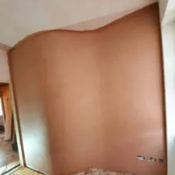 Świeżo nałożona, nierówna warstwa brązowej zaprawy na ścianie, widoczne pofalowania przy suficie, przygotowanie do dalszych prac wykończeniowych, zdemontowana podłoga i wystające kable elektryczne.