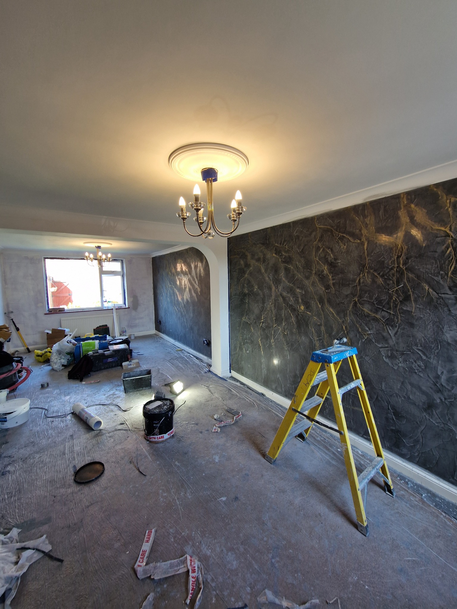 Pokój w trakcie remontu z dekoracyjną ścianą w odcieniach szarości i złota, żółta drabina, narzędzia malarskie i zabezpieczona folią podłoga.