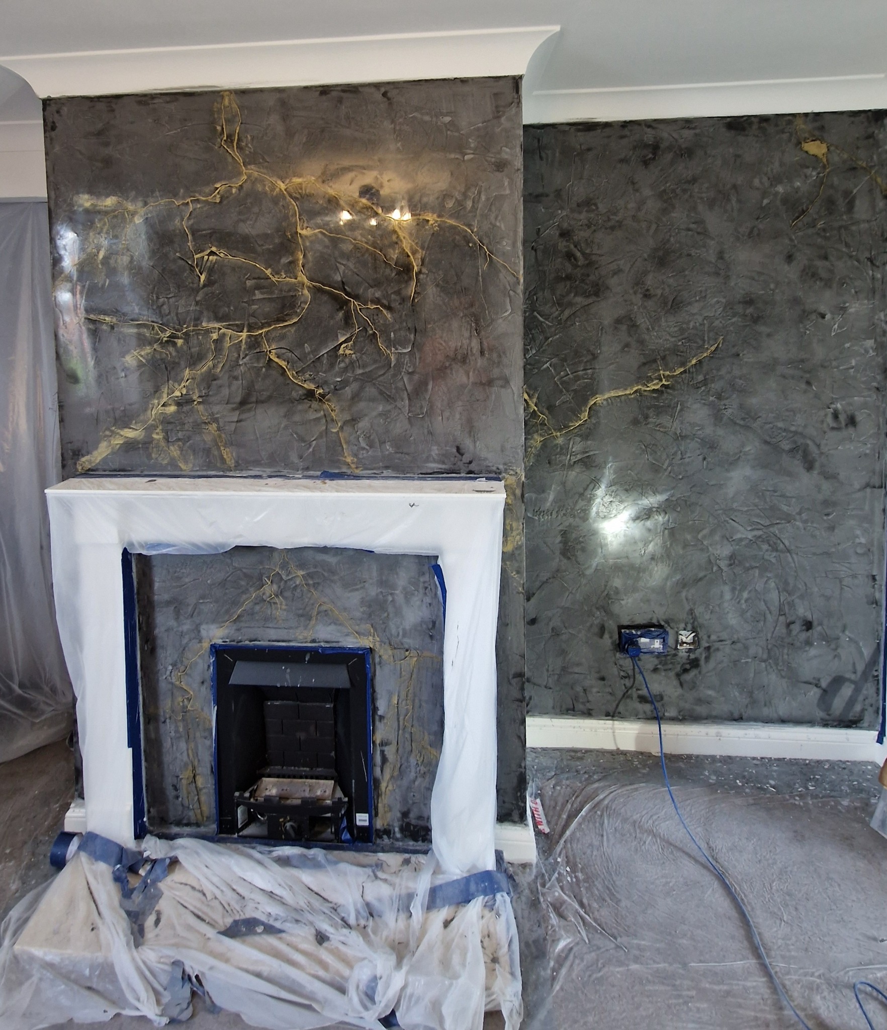 Pokój w trakcie prac wykończeniowych: ściana z dekoracyjną, ciemną fakturą z złotymi żyłkami, kominek zabezpieczony folią malarską, podłoga również zabezpieczona folią.