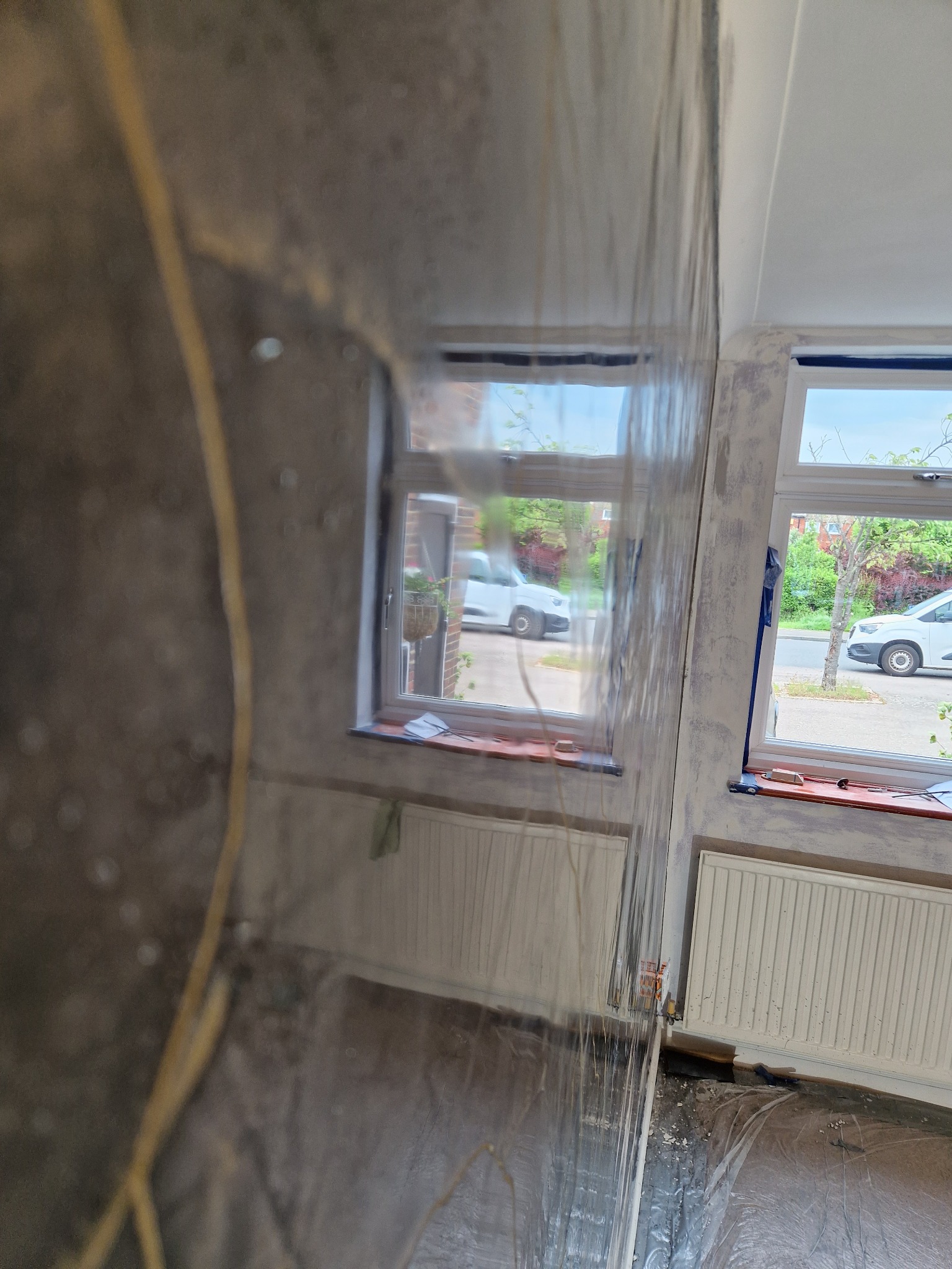 Przygotowanie do szpachlowania ścian w pokoju, folia ochronna na ścianie i podłodze, okna zabezpieczone taśmą malarską, widoczne grzejniki i fragment ściany przed nałożeniem gładzi.