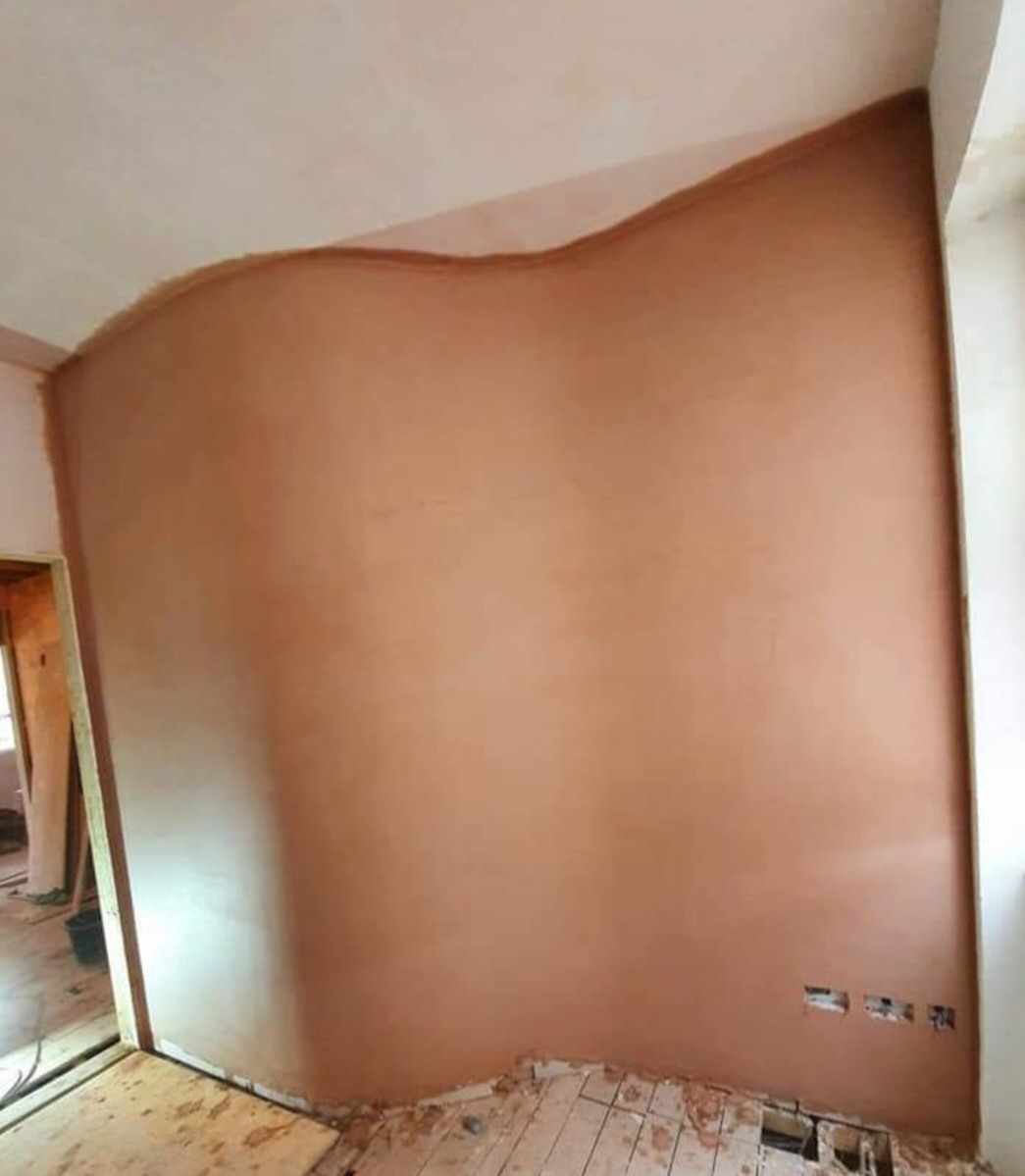 Świeżo nałożona, nierówna warstwa brązowej zaprawy na ścianie, widoczne pofalowania przy suficie, przygotowanie do dalszych prac wykończeniowych, zdemontowana podłoga i wystające kable elektryczne.