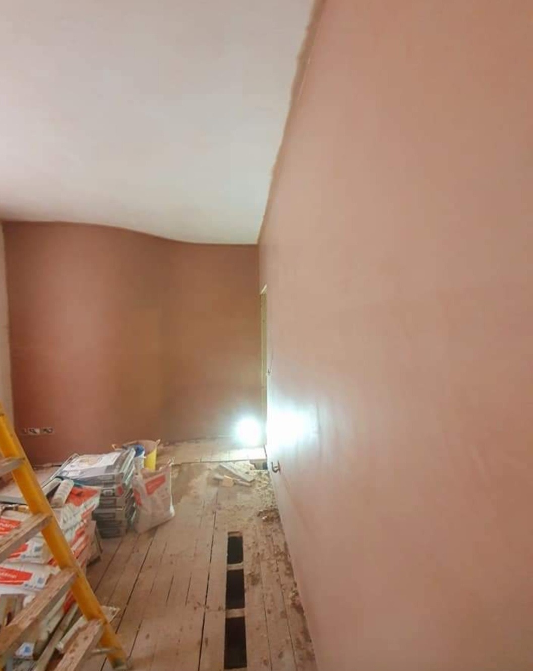 Świeżo otynkowane ściany w pomieszczeniu, widoczne worki z zaprawą i drabina, podłoga częściowo odkryta, przygotowana do dalszych prac.