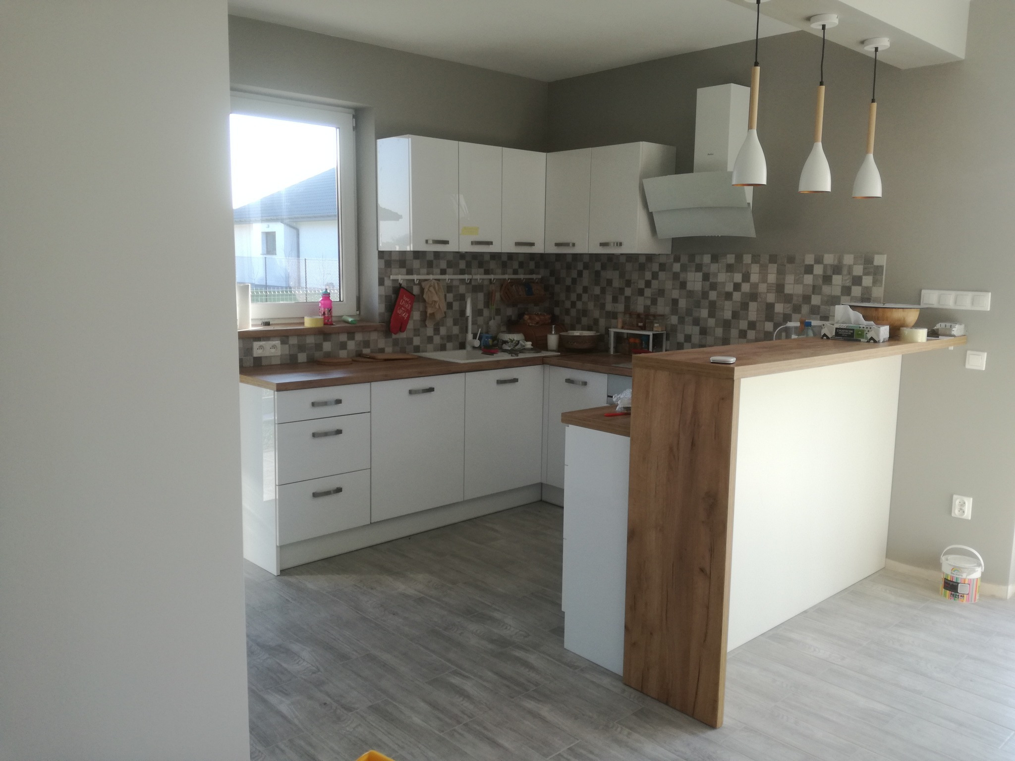 Nowoczesna kuchnia w odcieniach bieli i szarości z drewnianymi akcentami, widoczne wiszące lampy z drewnianymi elementami, mozaika nad blatem i wiaderko z farbą w rogu pomieszczenia.