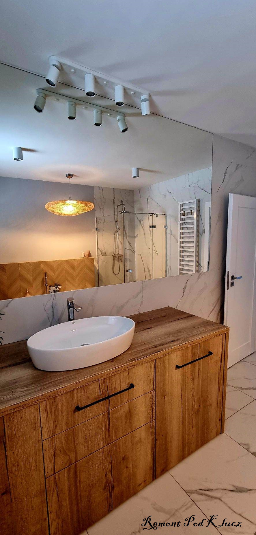 Nowoczesna łazienka po remoncie: owalna umywalka nablatowa na drewnianej szafce z czarnymi uchwytami, lustro na ścianie z marmurowym wzorem, widoczna kabina prysznicowa i grzejnik drabinkowy...