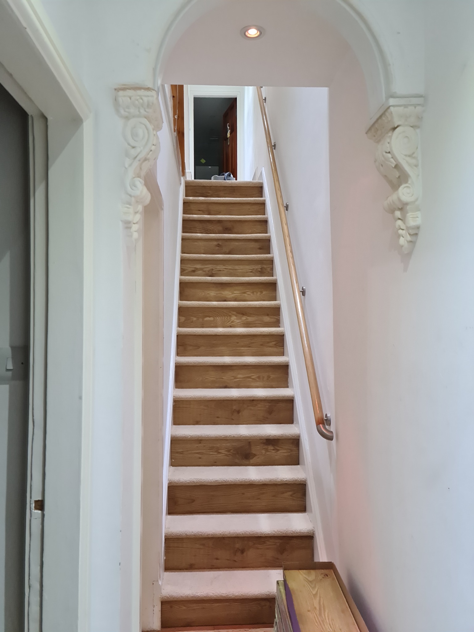 Schody drewniane z wykładziną dywanową, białe ściany z dekoracyjnymi wspornikami i drewnianą poręczą, widok z dołu.