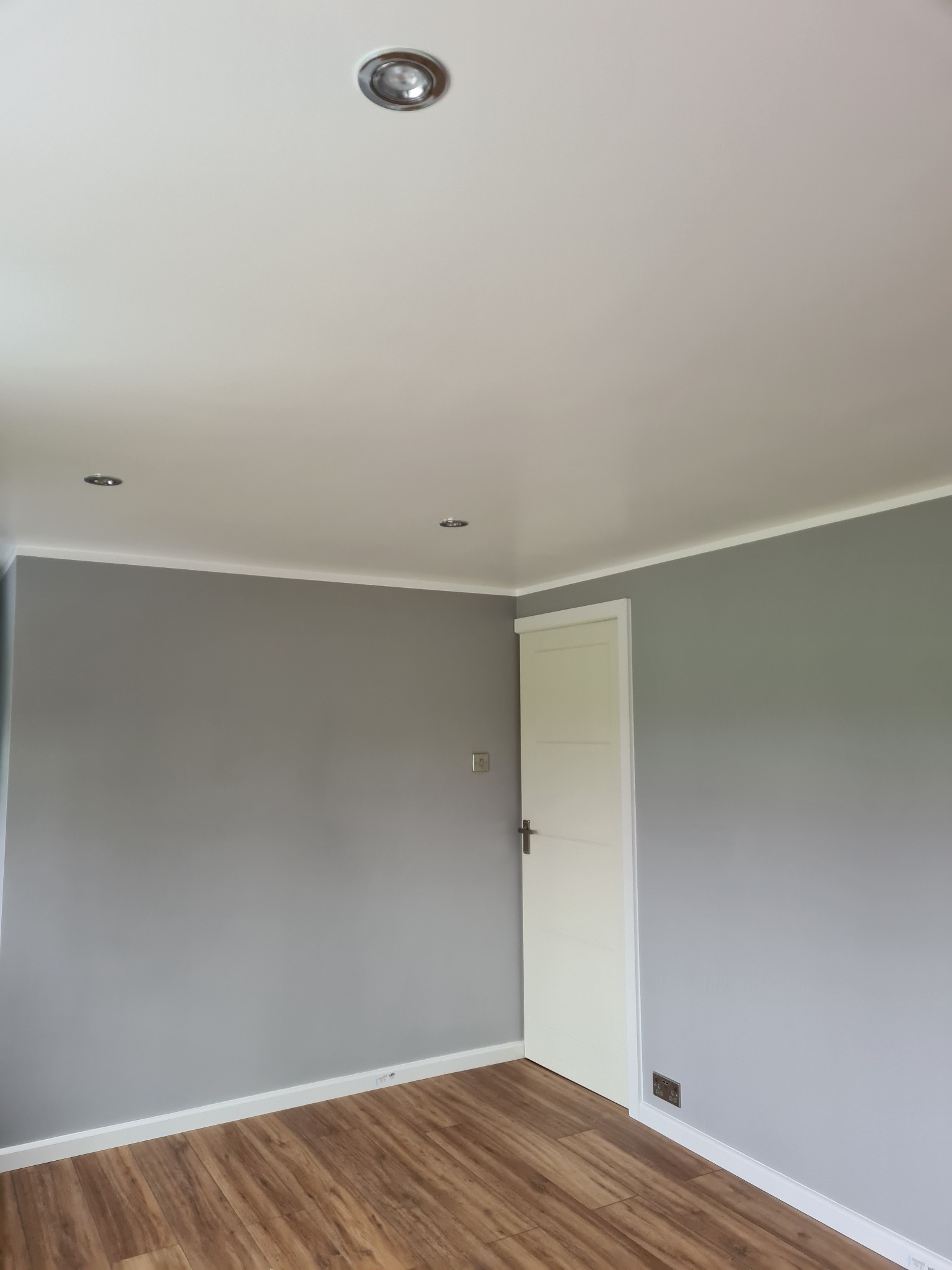 Pomalowane na szaro wnętrze pokoju z drewnianą podłogą, białymi listwami przypodłogowymi i sufitowymi oraz trzema punktowymi lampami w suficie.