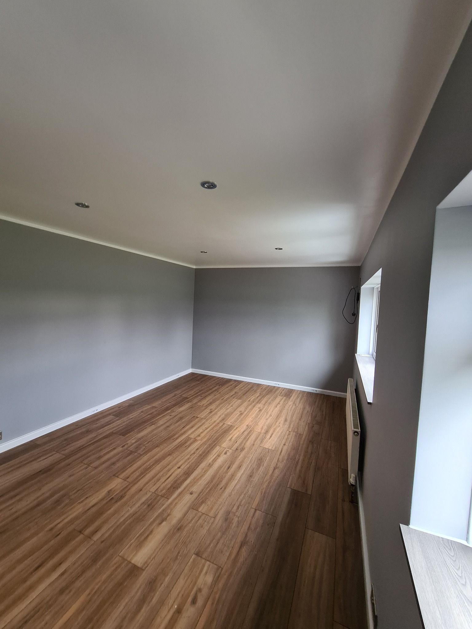 Pusty pokój po remoncie: szare ściany, drewnopodobna podłoga z paneli, biała listwa przypodłogowa i sufitowa, okno z grzejnikiem, widoczne kable przy oknie.