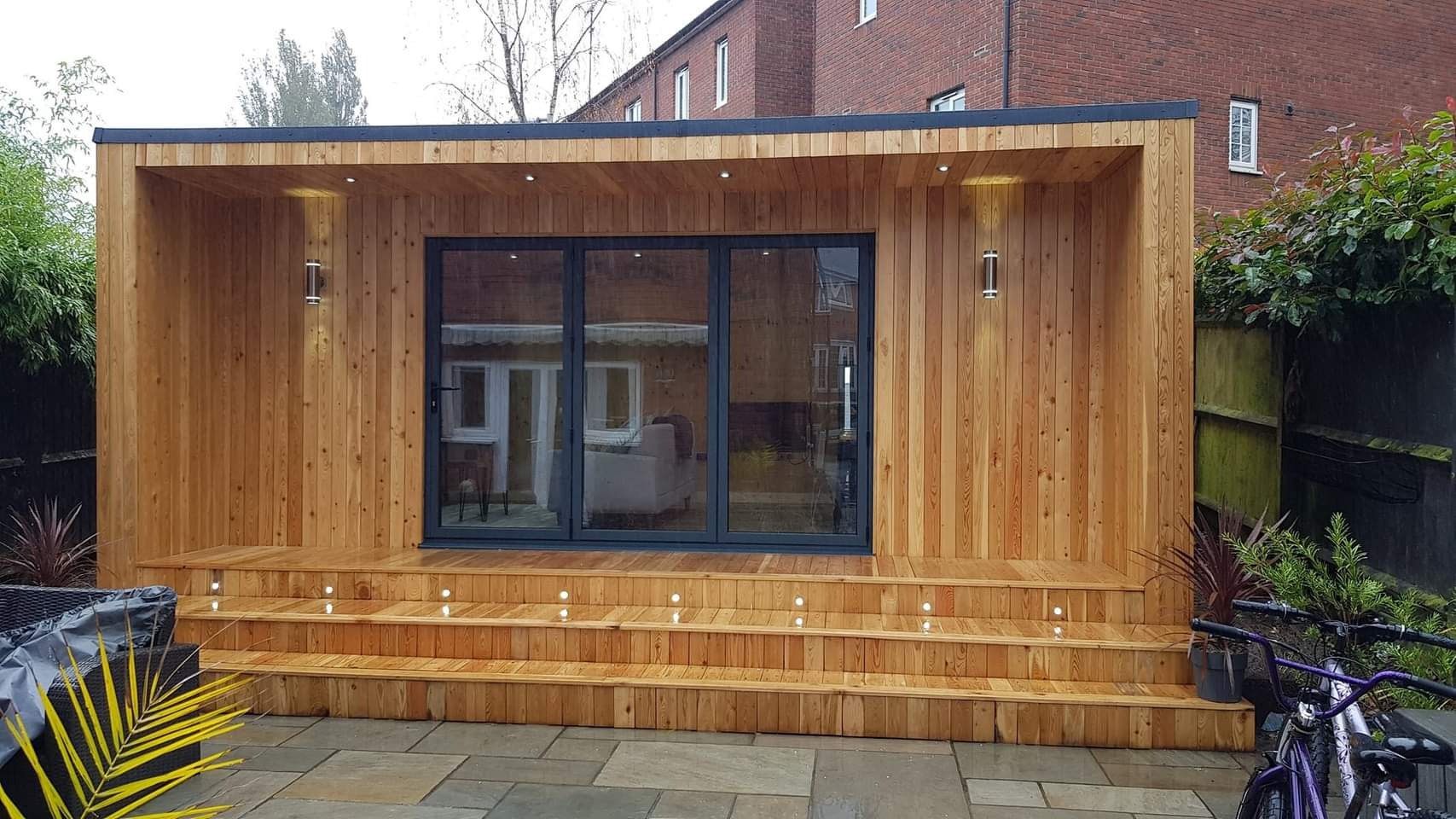 Nowoczesny drewniany pawilon ogrodowy z trzema schodkami z wbudowanym oświetleniem, szare, składane drzwi przesuwne, zewnętrzne lampy ścienne i kamienna ścieżka