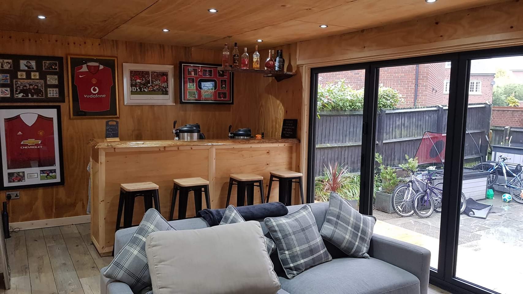 Drewniany bar w domowym pomieszczeniu z piłkarskimi pamiątkami na ścianach, widok na ogród przez duże okno, szara sofa z poduszkami w kratę.