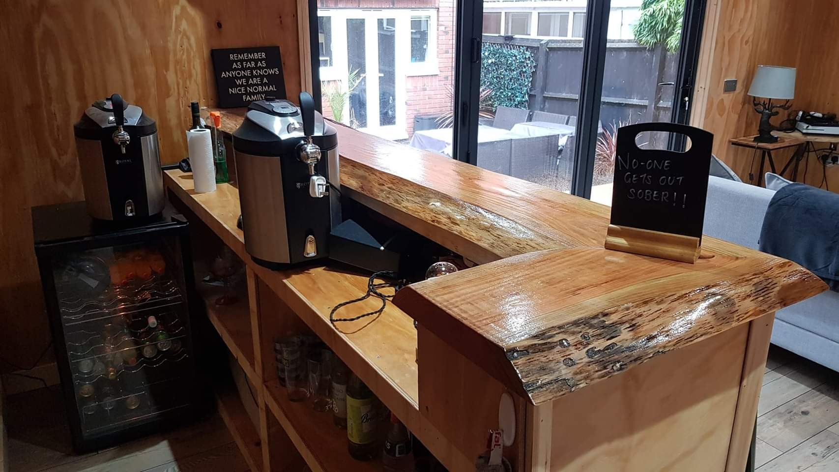 Drewniany bar z dwoma dystrybutorami do piwa, lodówką na napoje, tabliczką z napisem 'No-one gets out sober!!' i dekoracyjną tablicą z napisem 'Remember as far as anyone knows we are a nice normal...