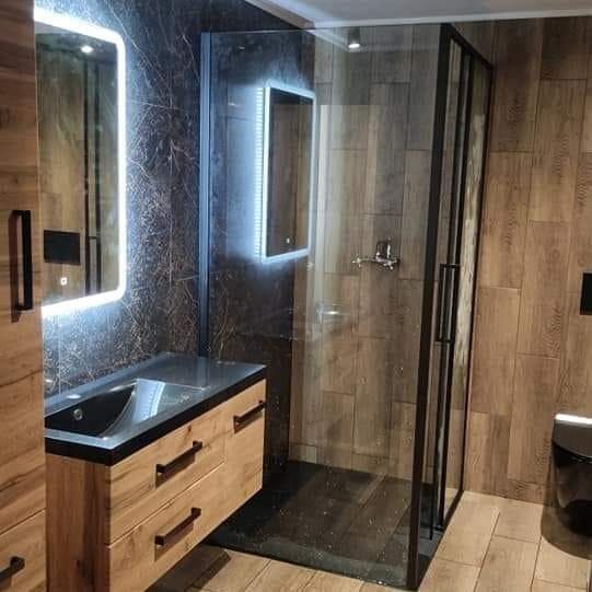 Nowoczesna łazienka z kabiną prysznicową z czarnymi profilami, drewnopodobnymi płytkami i podświetlanymi lustrami.