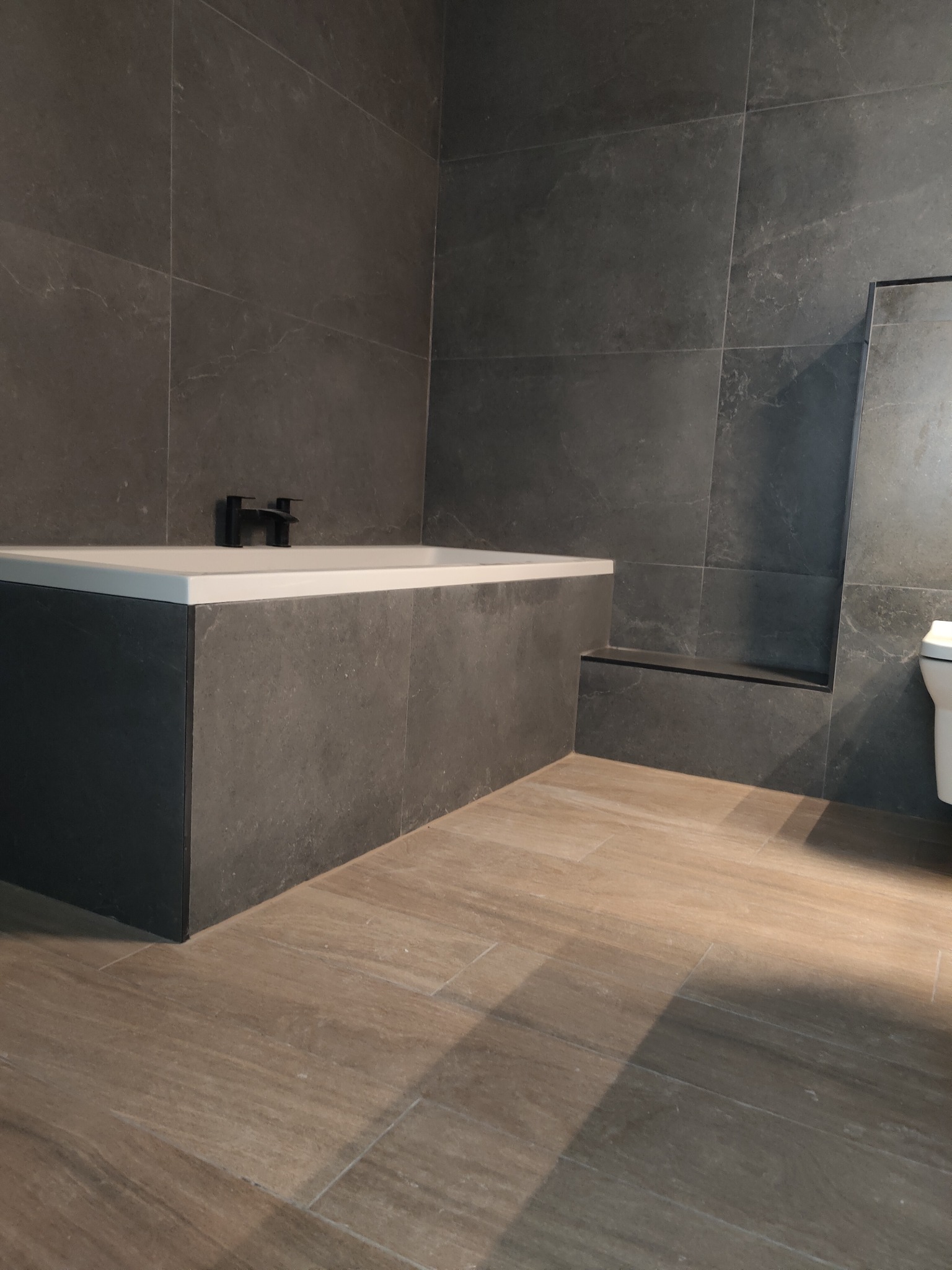 Nowoczesna łazienka z ciemnymi płytkami imitującymi kamień na ścianach i obudowie wanny, drewnopodobne płytki na podłodze, czarna bateria wannowa.