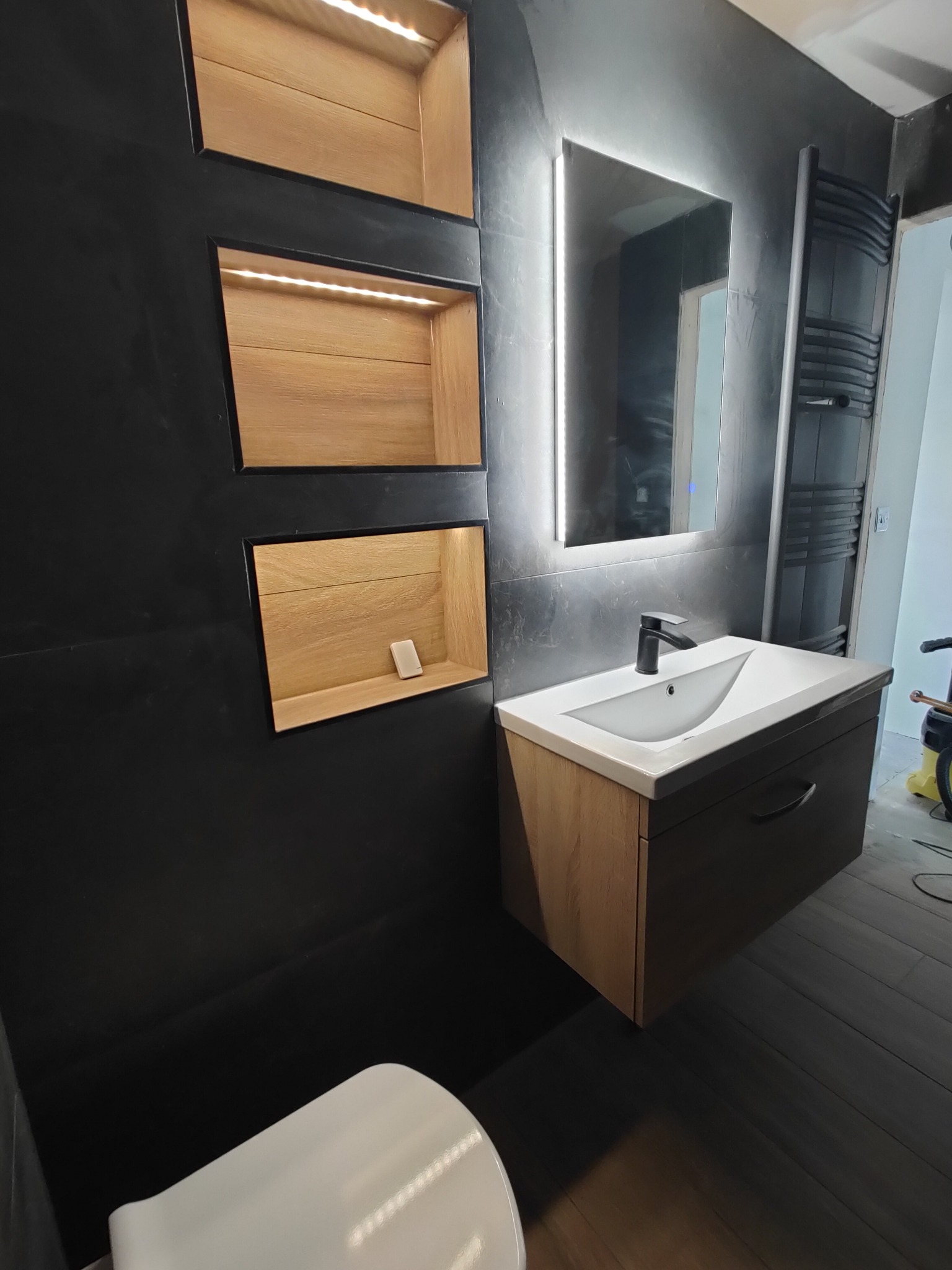 Nowoczesna łazienka z czarnymi ścianami, drewnianymi wnękami z oświetleniem LED, podświetlanym lustrem i umywalką z szafką w odcieniach brązu i szarości.